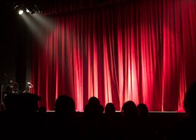 Dimly lit theater stage with red curtains and audience silhouettes under spotlights.